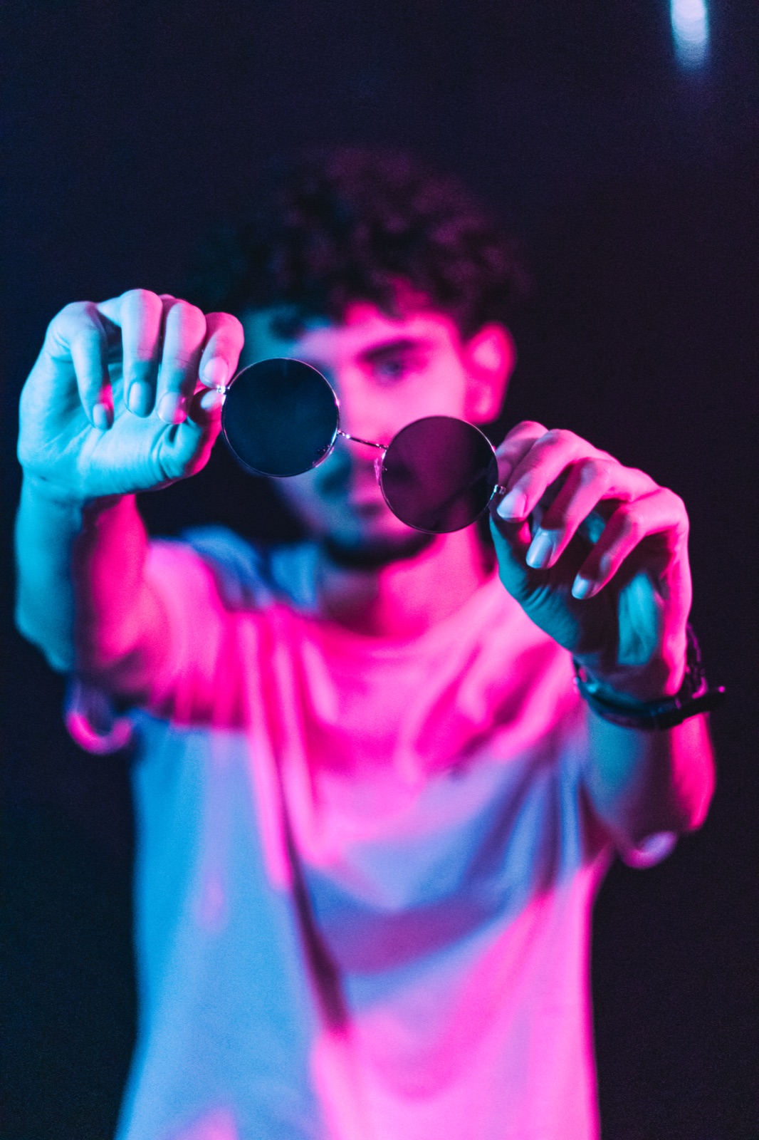 Neon-lit studio portrait, round sunglasses held out toward the camera